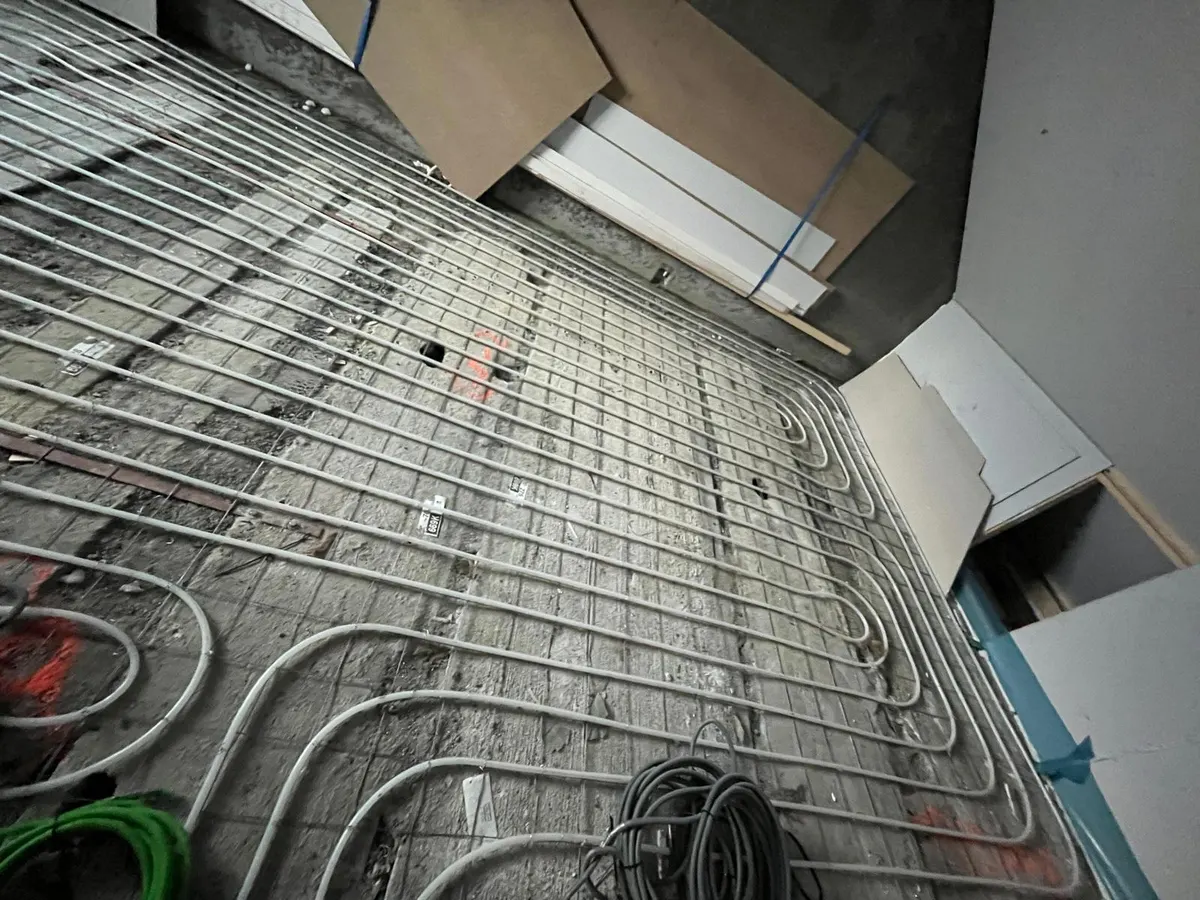 Een wapeningsnet met vloerverwarmingsbuis in dichte banen van dichtbij, met opgerolde kabels op de betonvloer en houten en kartonnen platen tegen de wand erachter