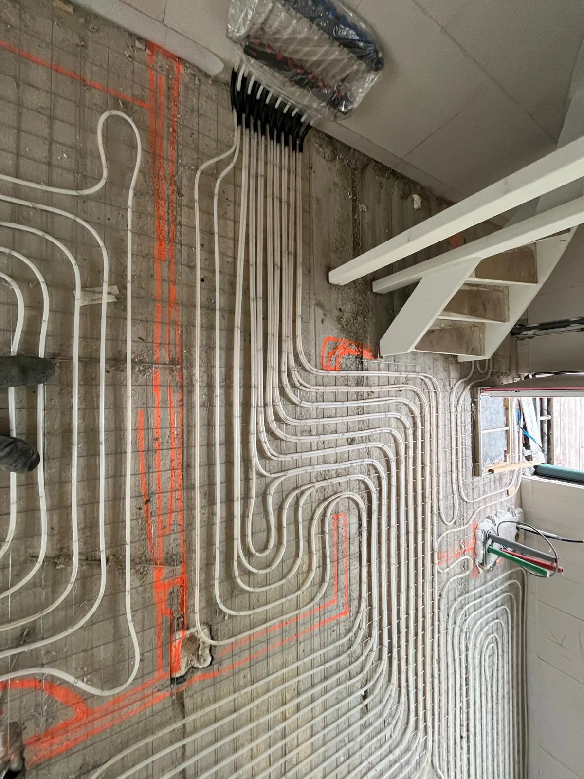 Vloerverwarmingsbuis in ruime lussen op een wapeningsnet op een ruwe betonvloer, met oranje uitzetlijnen, een witte trap rechts en een bundel zwarte leidingen langs de wand