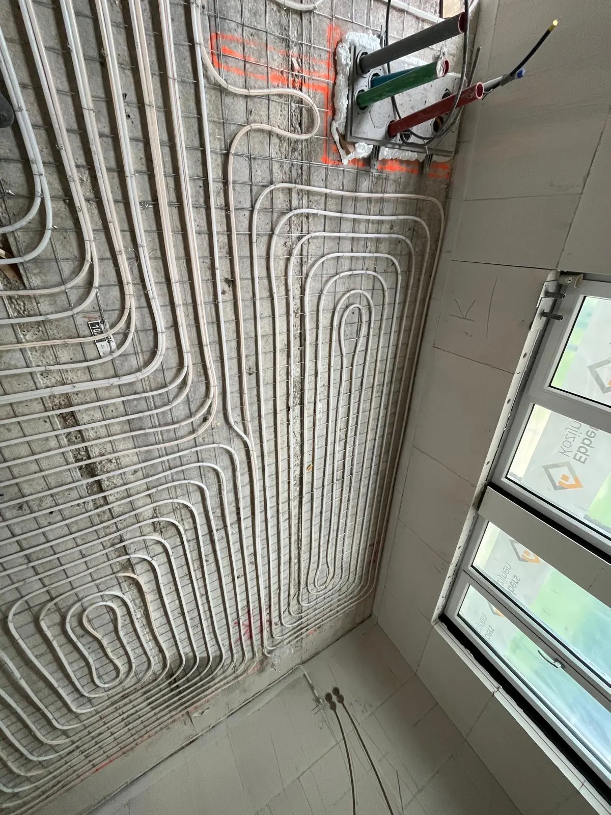 De hoek van een kamer met vloerverwarmingsbuis in lussen op een wapeningsnet, met een raamkozijn met beschermfolie rechts en oranje uitzetlijnen op de vloer