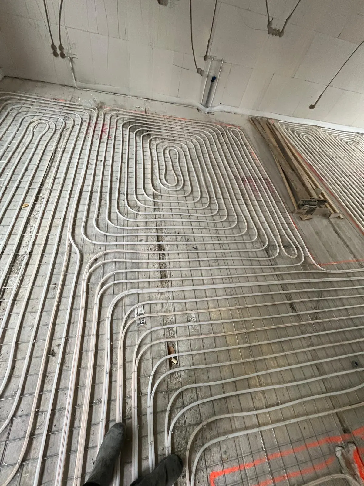 Een kamervloer vol vloerverwarmingsbuis op een wapeningsnet, met een stapel houten latten tegen de wand en de kale betonvloer langs de rand