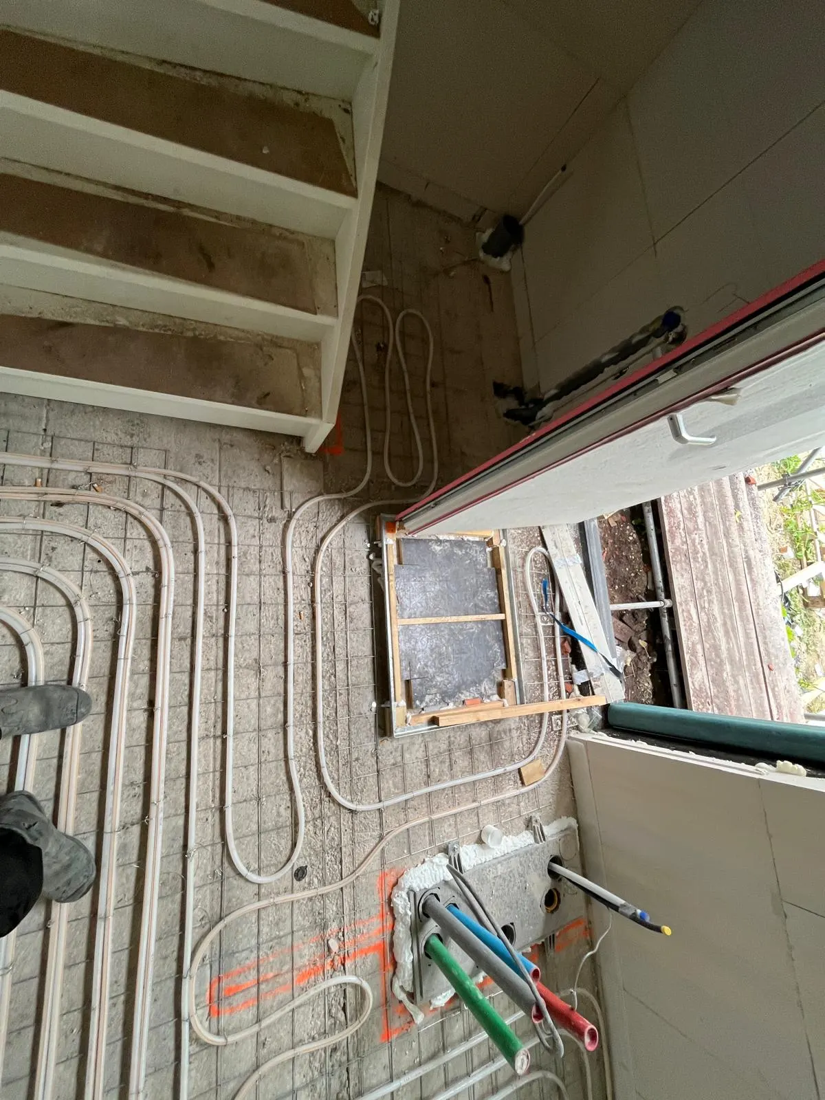 Vloerverwarmingsbuis om een muuropening heen gelegd in een klein vertrek, met een houten raamsponning in de wand en groene en rode aanvoerleidingen die uit de wand steken