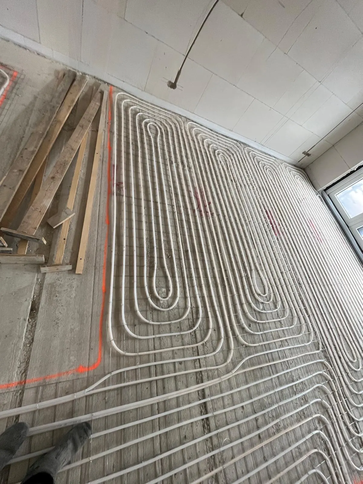 Een langgerekte kamer met vloerverwarmingsbuis in lange lussen op een wapeningsnet, met een stapel houten latten links en een raam aan het eind