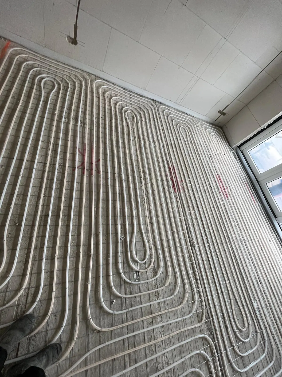 Vloerverwarmingsbuis in lange banen op een wapeningsnet over een hele kamervloer, met twee ramen in de wand rechts