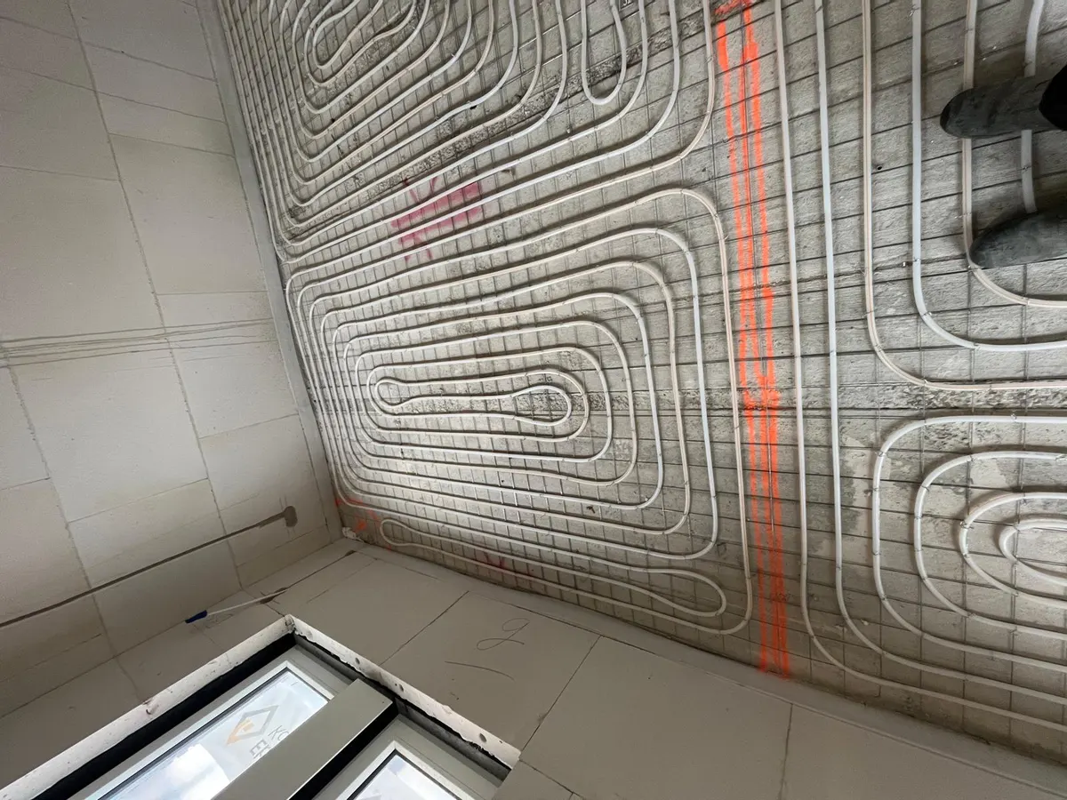 Een kamerhoek met vloerverwarmingsbuis in lussen op een wapeningsnet, met een oranje uitzetlijn dwars over de vloer en twee ramen laag in de wand