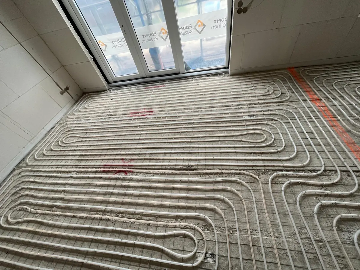 Een vloer vol vloerverwarmingsbuis op een wapeningsnet tot aan een schuifpui met beschermfolie, met de kale betonrand langs de wand