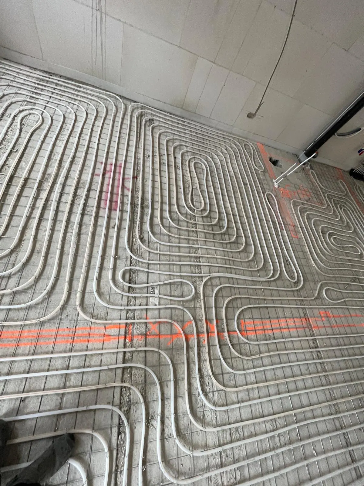 Vloerverwarmingsbuis in ineengeschoven lussen op een wapeningsnet, met oranje uitzetlijnen kruislings over de vloer en een leiding tegen de wand