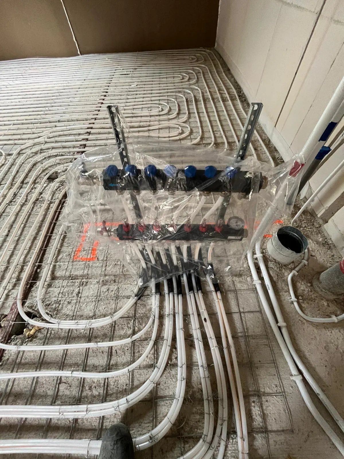 De verdeler van de woning in beschermfolie aan de wand boven de vloer, met de aansluitleidingen die er in een waaier naartoe lopen en een grijze standleiding ernaast
