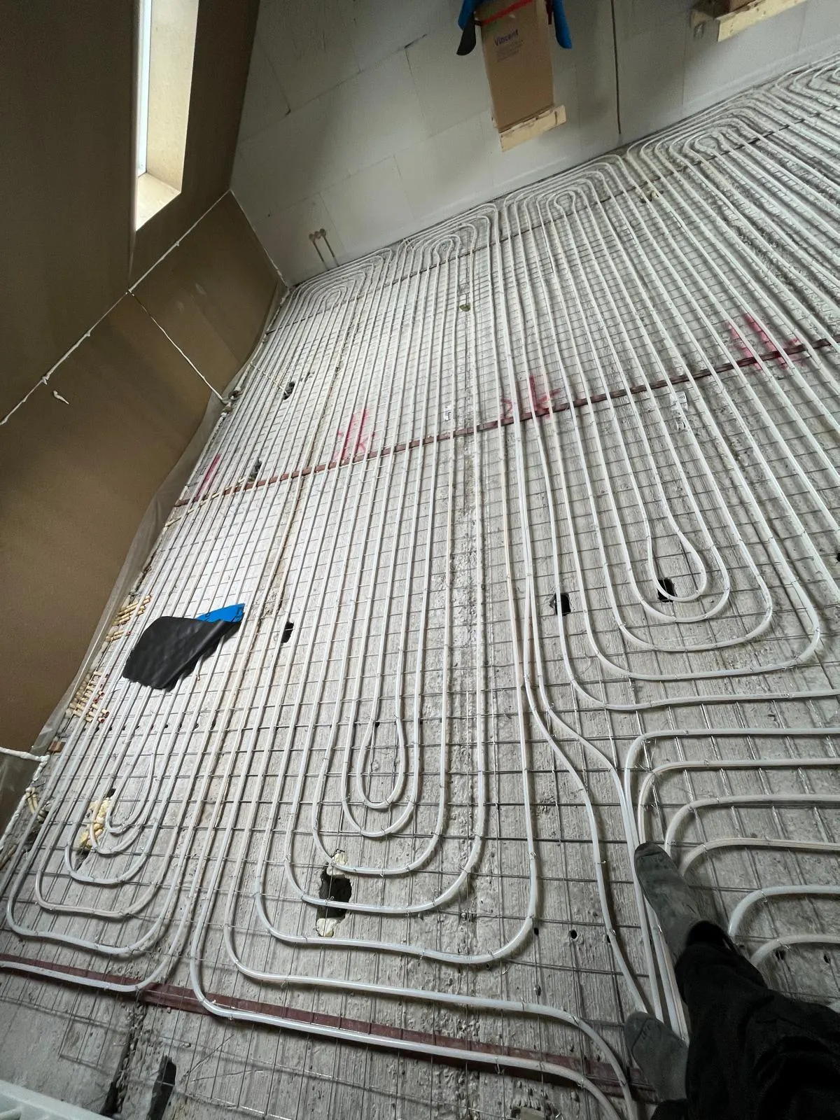 Een zolderkamer met vloerverwarmingsbuis op een wapeningsnet tot onder de schuine wand, met een smal raam links en werkspullen op de vloer