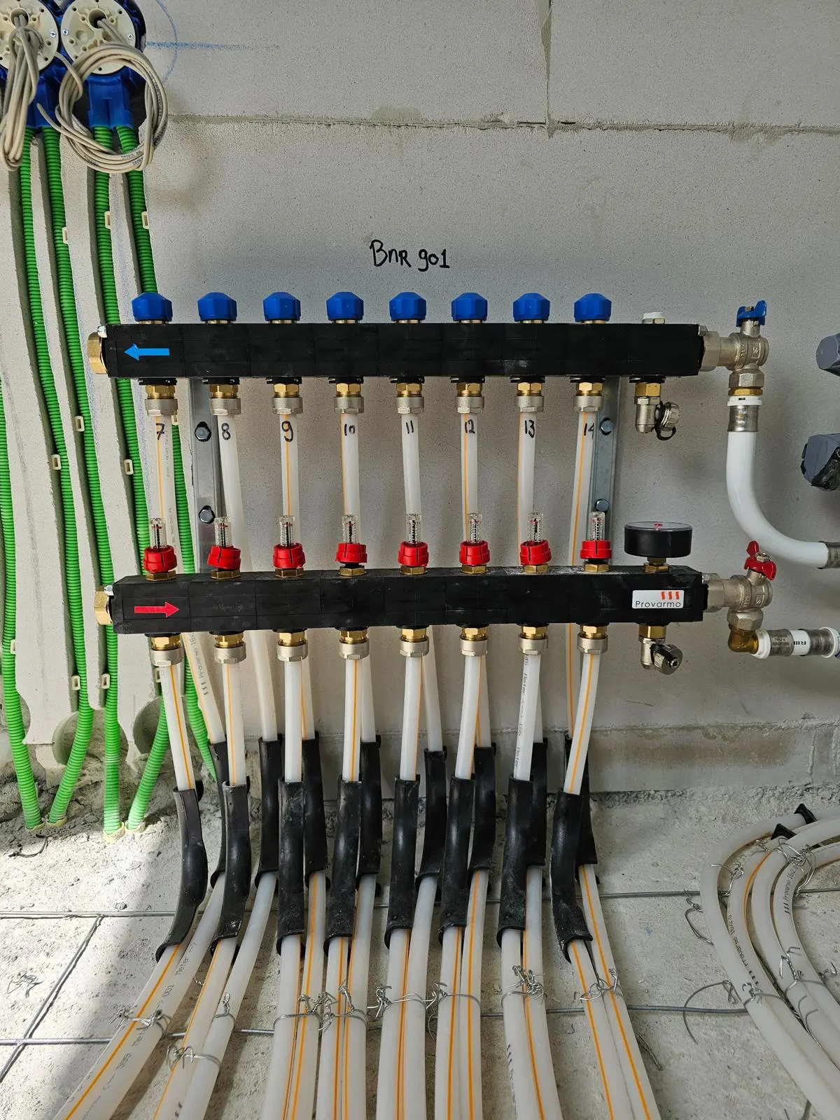 Een verdeler met acht genummerde groepen aan een kalkzandsteenwand, met blauwe afsluiters op de bovenste rail, rode op de onderste, een manometer en groene elektrabuizen ernaast