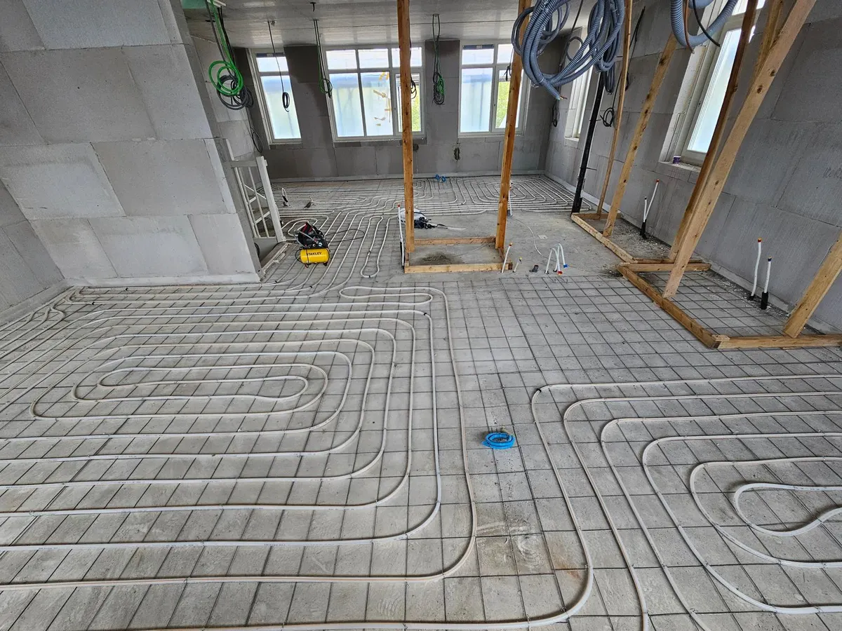 Een grote open verdiepingsvloer in ruwbouw vol vloerverwarmingsbuis in lussen op een wapeningsnet, met kalkzandsteenwanden, houten stempels en een rij ramen op de achtergrond