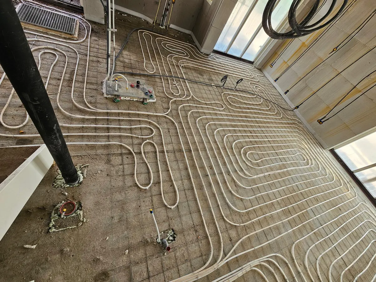 Vloerverwarmingsbuis in brede lussen en een slakkenhuis op een wapeningsnet over een ruwe vloer, met een zwarte standleiding en ingegoten doorvoeren links en een glazen pui rechts