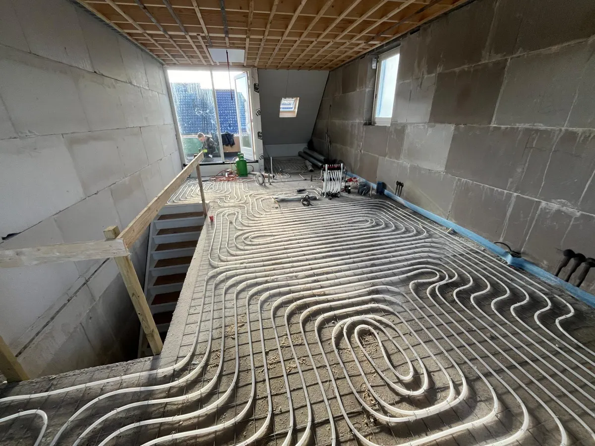 Een zolder met vloerverwarmingsbuis in ruime lussen op een wapeningsnet over de volle vloer, met een houten balklaag als plafond, een trapgat met een houten hekwerk links en daglicht uit een balkondeur aan het eind