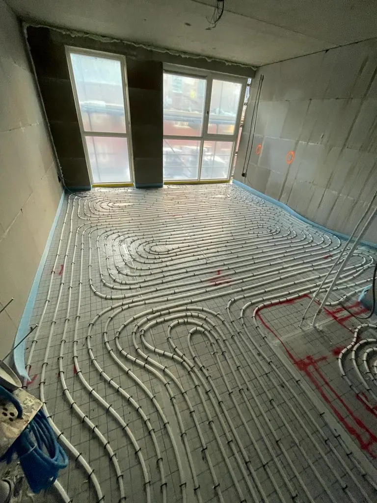 Vloerverwarmingsbuis in dichte lussen op een wapeningsnet in een appartement, met rode uitzetmarkeringen op de vloer en op de wand, blauwe randisolatie rondom en een pui met daglicht