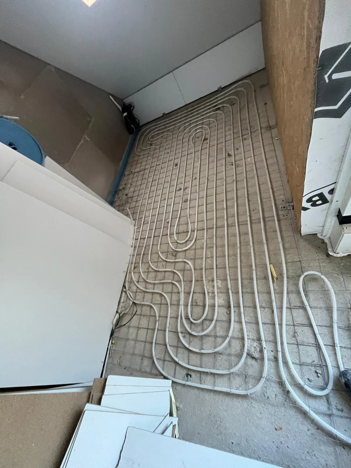 Vloerverwarmingsbuis in een lange lus op een wapeningsnet over een kamervloer, met gipsplaten tegen de wand en een blauwe haspel in de hoek