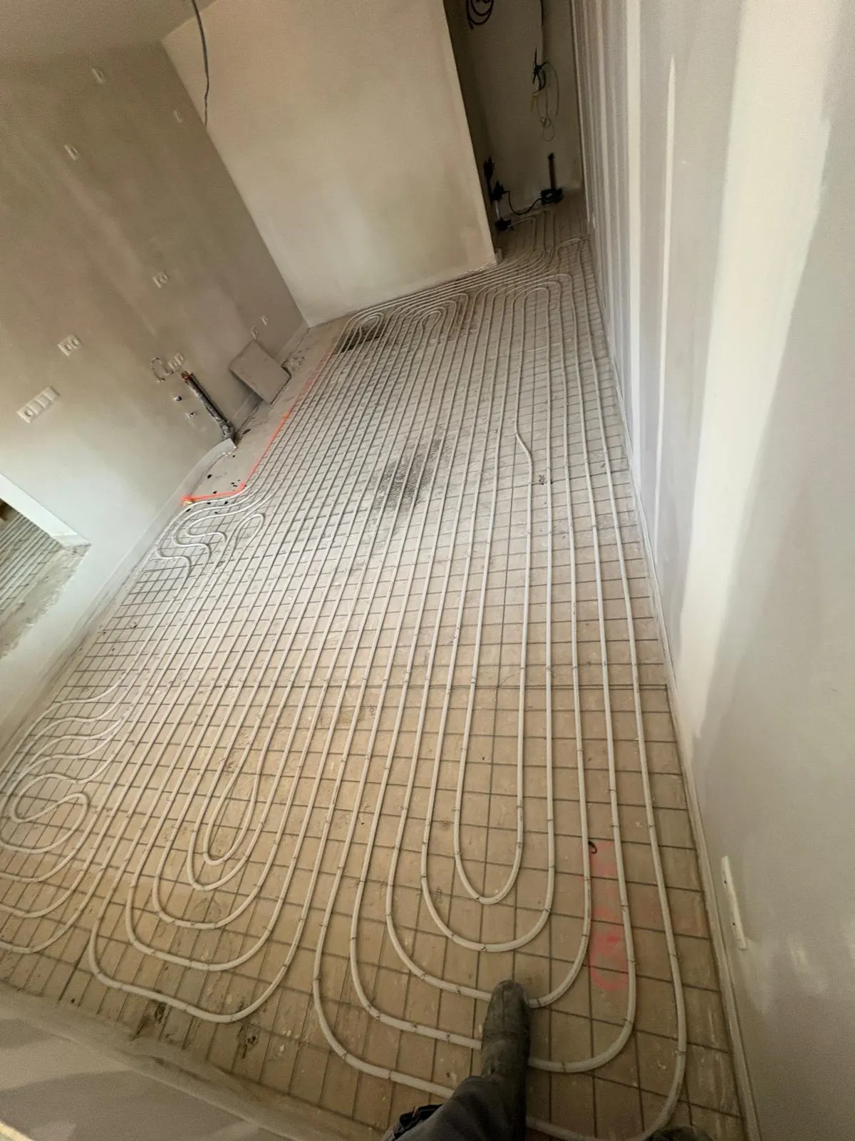 Vloerverwarmingsbuis in dichte lussen over de volle vloer van een smalle ruimte, op een wapeningsnet over lichtbruine plaatisolatie, met een oranje uitzetlijn langs de wand en een aftakking die door een deuropening de gang in loopt
