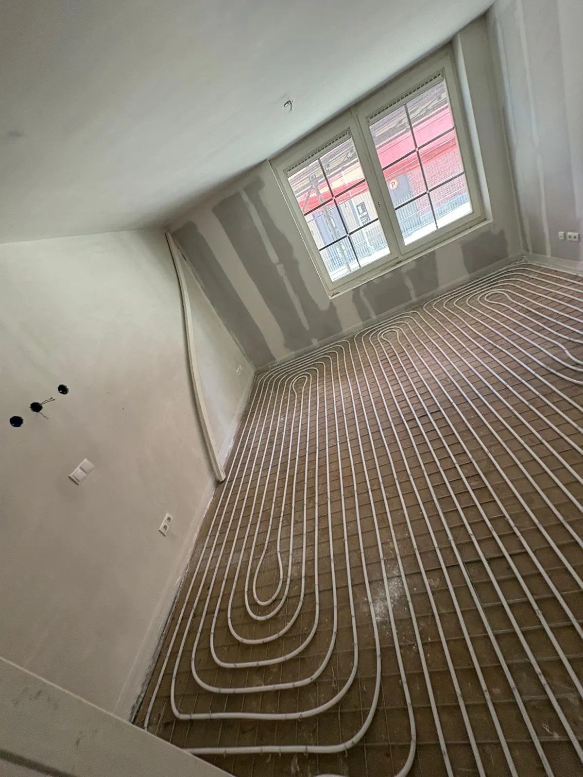 Een kamer onder de kap met een schuine wand, met vloerverwarmingsbuis in dichte lussen op een wapeningsnet en een kozijn met roedes waardoor de rode baksteengevel aan de overkant te zien is