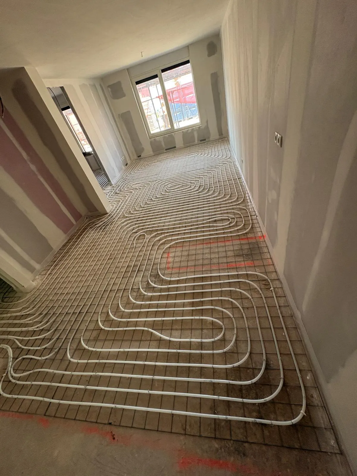 Een langgerekte ruimte met twee deuropeningen op een rij, met vloerverwarmingsbuis in ruime bochten en een slakkenhuis in het midden, en steigerwerk achter het raam