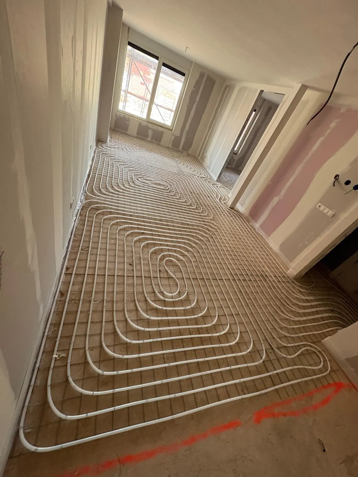 Vloerverwarmingsbuis in een slakkenhuis midden op een kamervloer, met een roze markeringslijn op het kale beton op de voorgrond en een doorgang rechts naar een ruimte met een geplamuurde wand