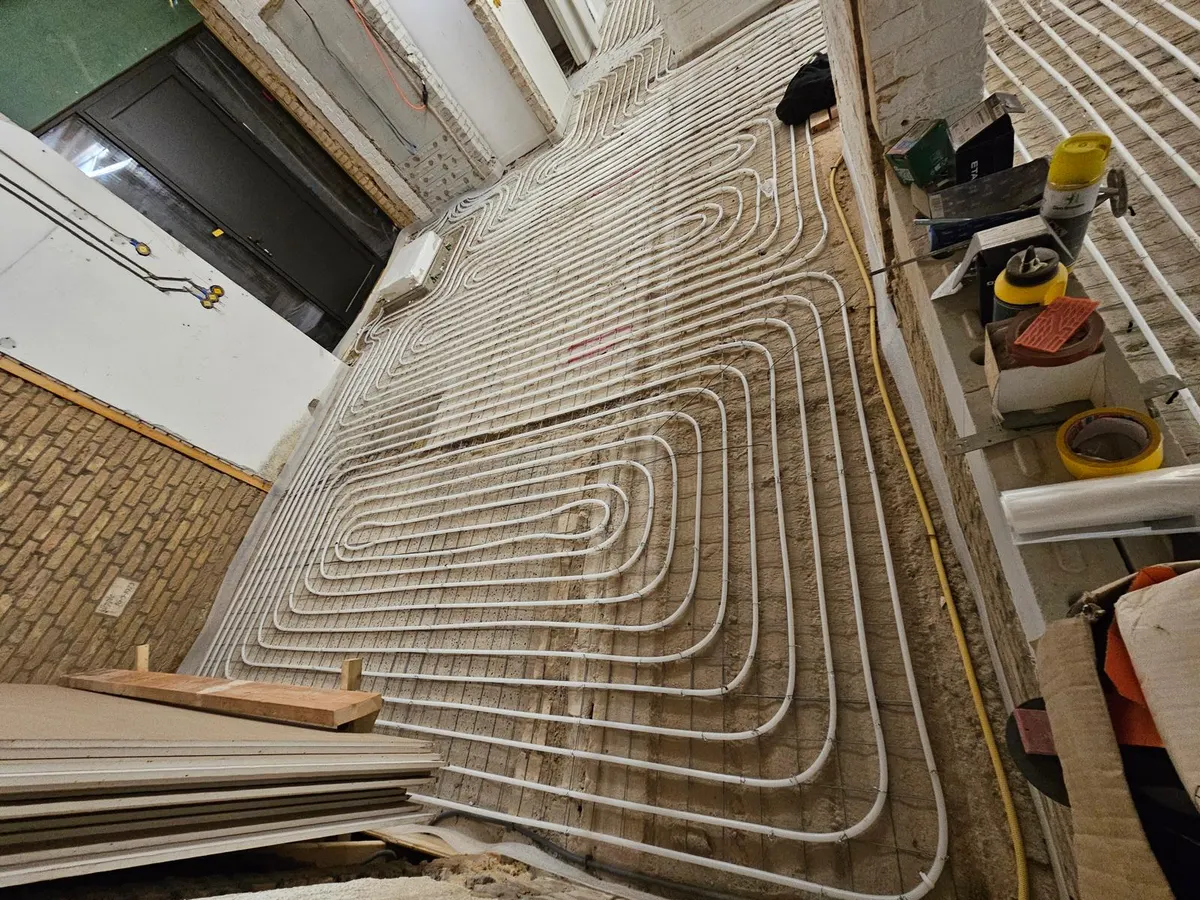 Vloerverwarmingsbuis in strakke lussen op een wapeningsnet over een vloer in ruwbouw, met een bakstenen wand, een deurpost en gereedschap op een werkbank langs de rand