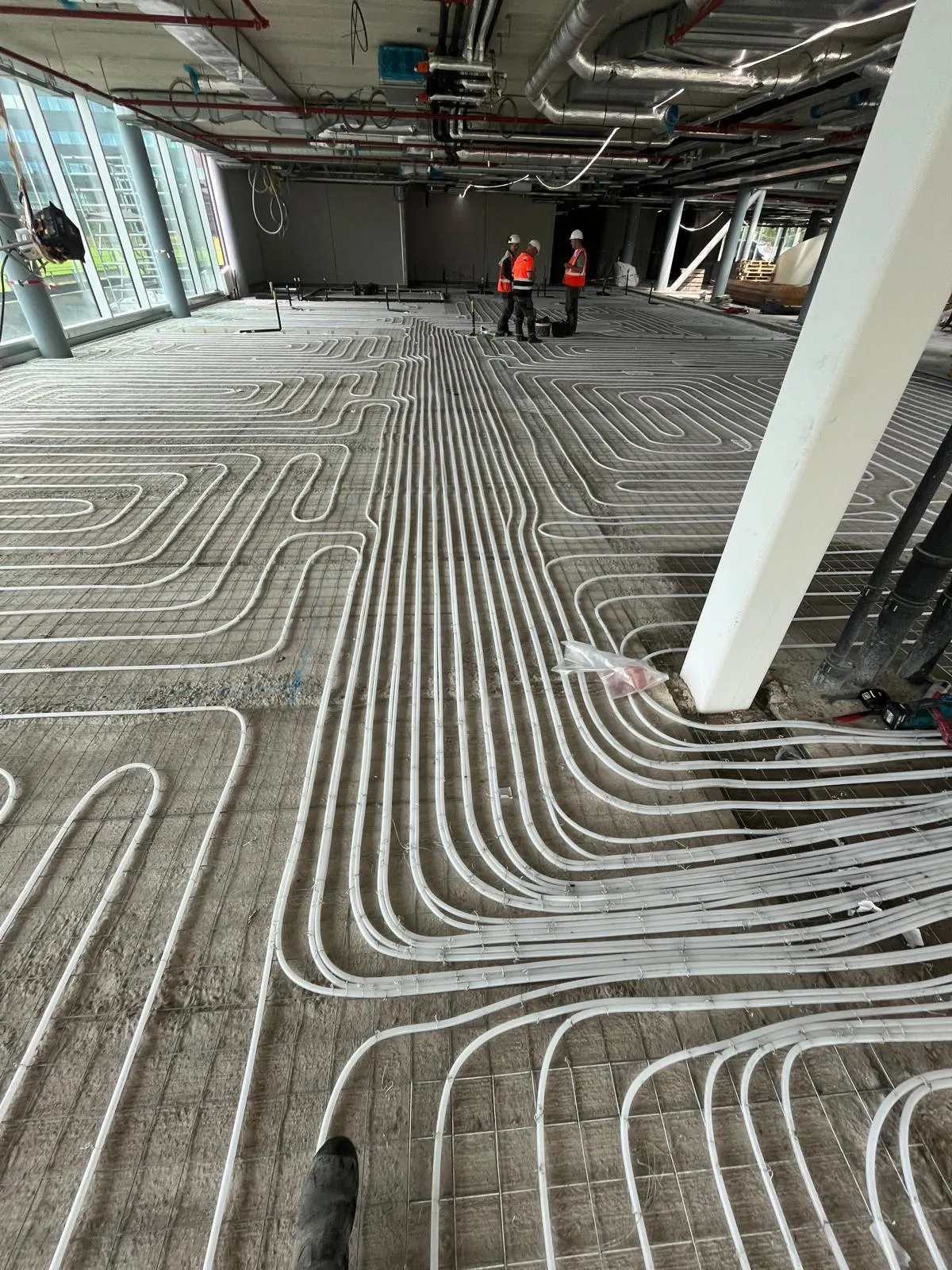 Een ruime verdiepingsvloer vol vloerverwarmingsbuis op een wapeningsnet, met een betonkolom in beeld, installatieleidingen aan het plafond en drie monteurs verderop