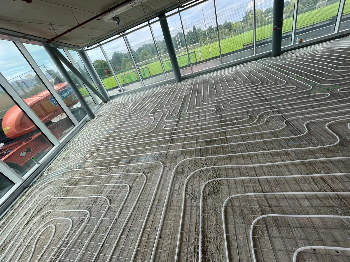 Vloerverwarmingsbuis in brede lussen op een wapeningsnet langs een glazen gevel met uitzicht op sportvelden