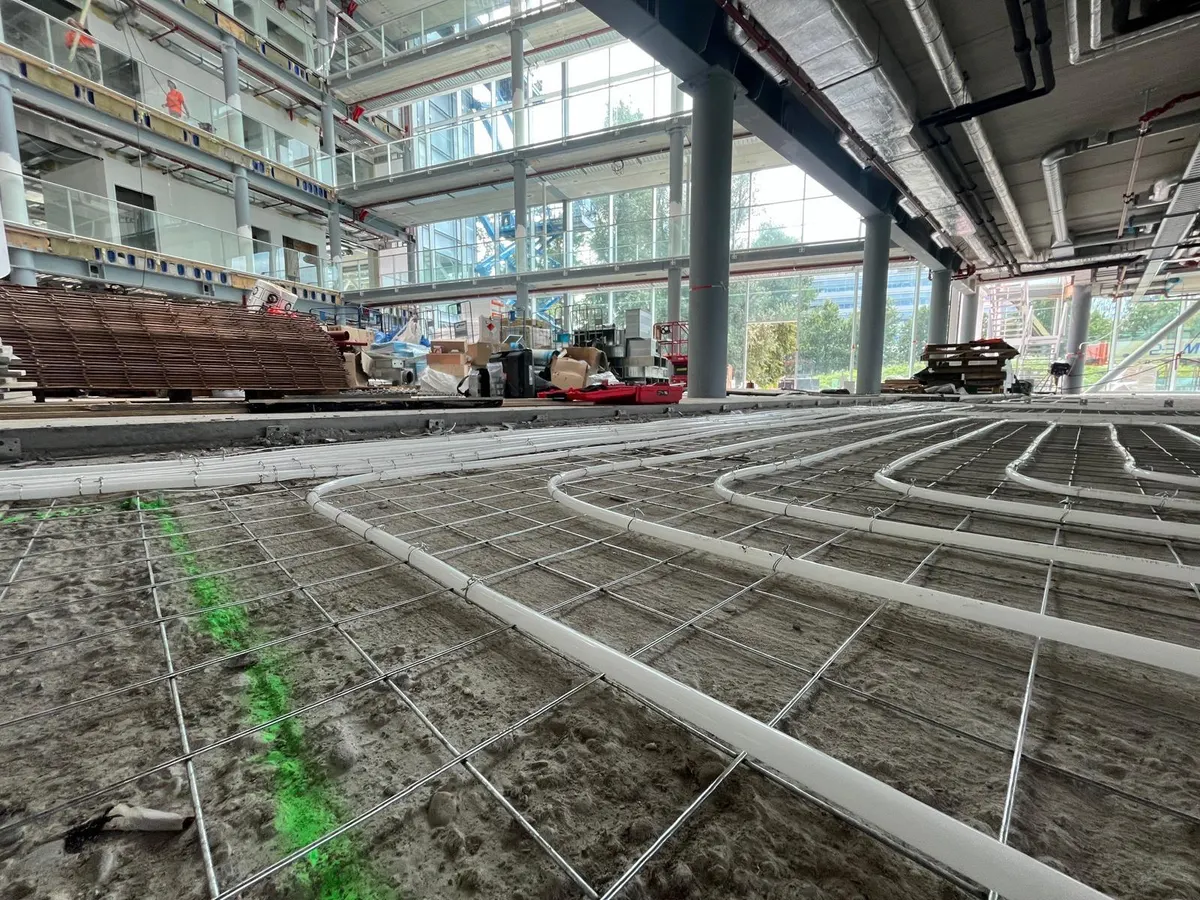 Vloerverwarmingsbuis op een wapeningsnet in een hoge hal met glazen gevels en galerijen erboven, met stapels wapeningsnet en materiaal langs de rand