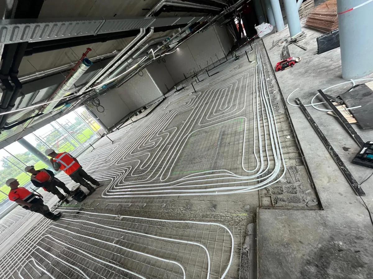 Een grote verdiepingsvloer vol vloerverwarmingsbuis in brede lussen op een wapeningsnet, met betonkolommen, installatieleidingen aan het plafond en drie monteurs bij de glazen gevel