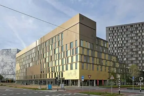 Een gevelfoto van een opgeleverd universiteitsgebouw van zes lagen met een goudkleurige gevel en smalle raamstroken, met de glazen entree op de begane grond en een kruispunt ervoor
