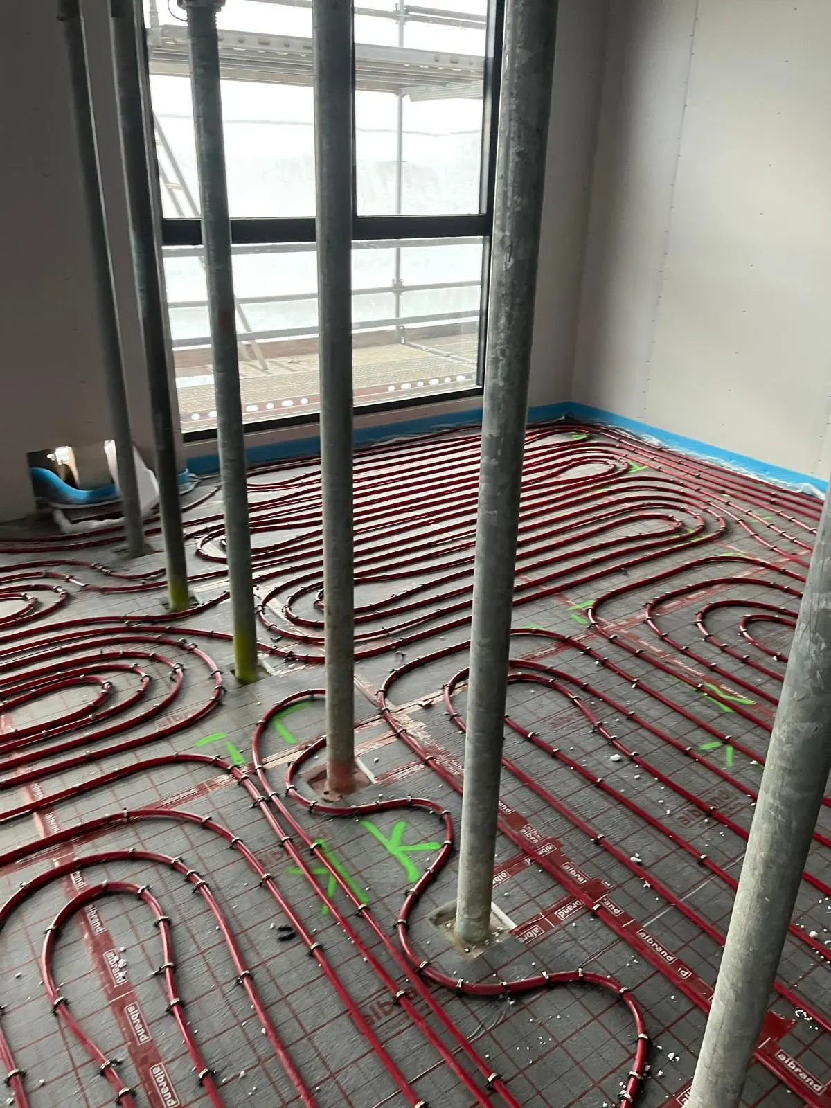 Een ruimte met een rij stempels onder het plafond, met rode vloerverwarmingsbuis in ruime bochten om de stempelvoeten heen gelegd, groene markeringen op de folie en een balkon achter de pui