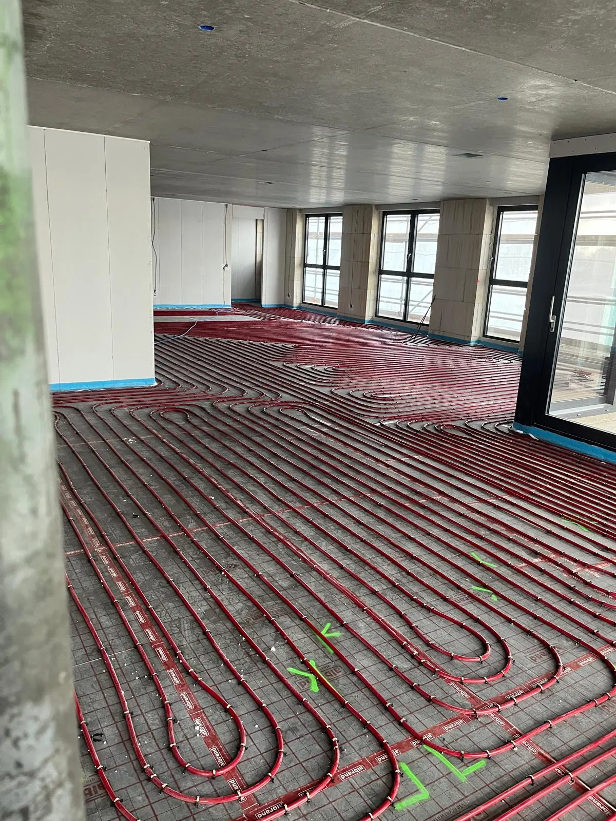 Een diep doorlopende verdiepingsvloer met een kale betonnen onderkant, met rode vloerverwarmingsbuis over de volle diepte tot tegen de raampartij, een scheidingswand links en een openslaande deur rechts