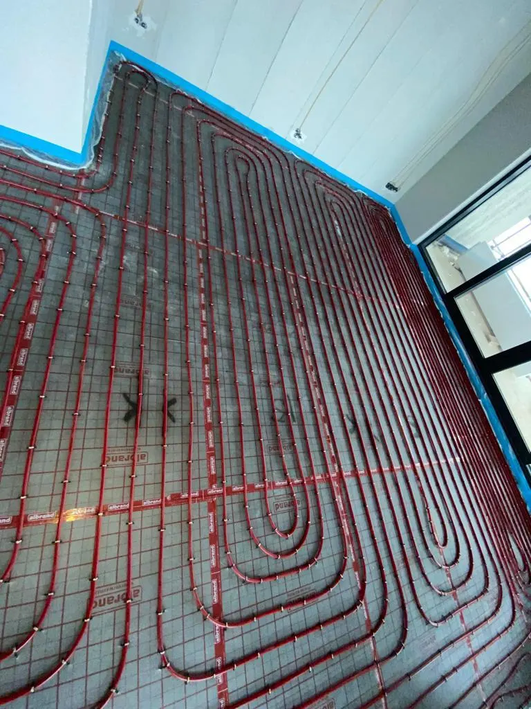 Rode vloerverwarmingsbuis in dichte lussen op tackerfolie over een hele kamervloer, met blauwe randisolatie langs de wanden en een donker raamkozijn rechts