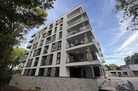 Een opgeleverd appartementengebouw van zes lagen in wit stucwerk met donkere raampartijen en balkons met glazen borstweringen, op een plint van natuursteen, met bomen ervoor en een blauwe lucht erboven