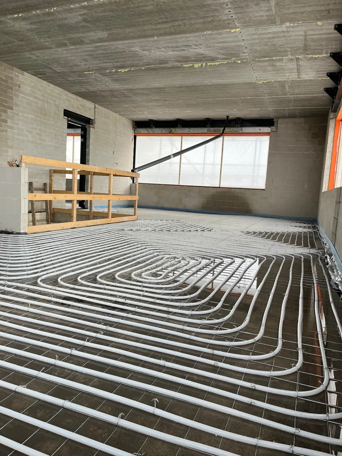 Een ruimte in ruwbouw met witte vloerverwarmingsbuis in dichte lussen, met bindijzer vastgezet op een wapeningsnet over een ruwe ondergrond, met kalkzandsteen wanden, een betonnen plafond, blauwe randisolatie langs de wand, een houten stelwand links en een groot raam met bouwnet erachter