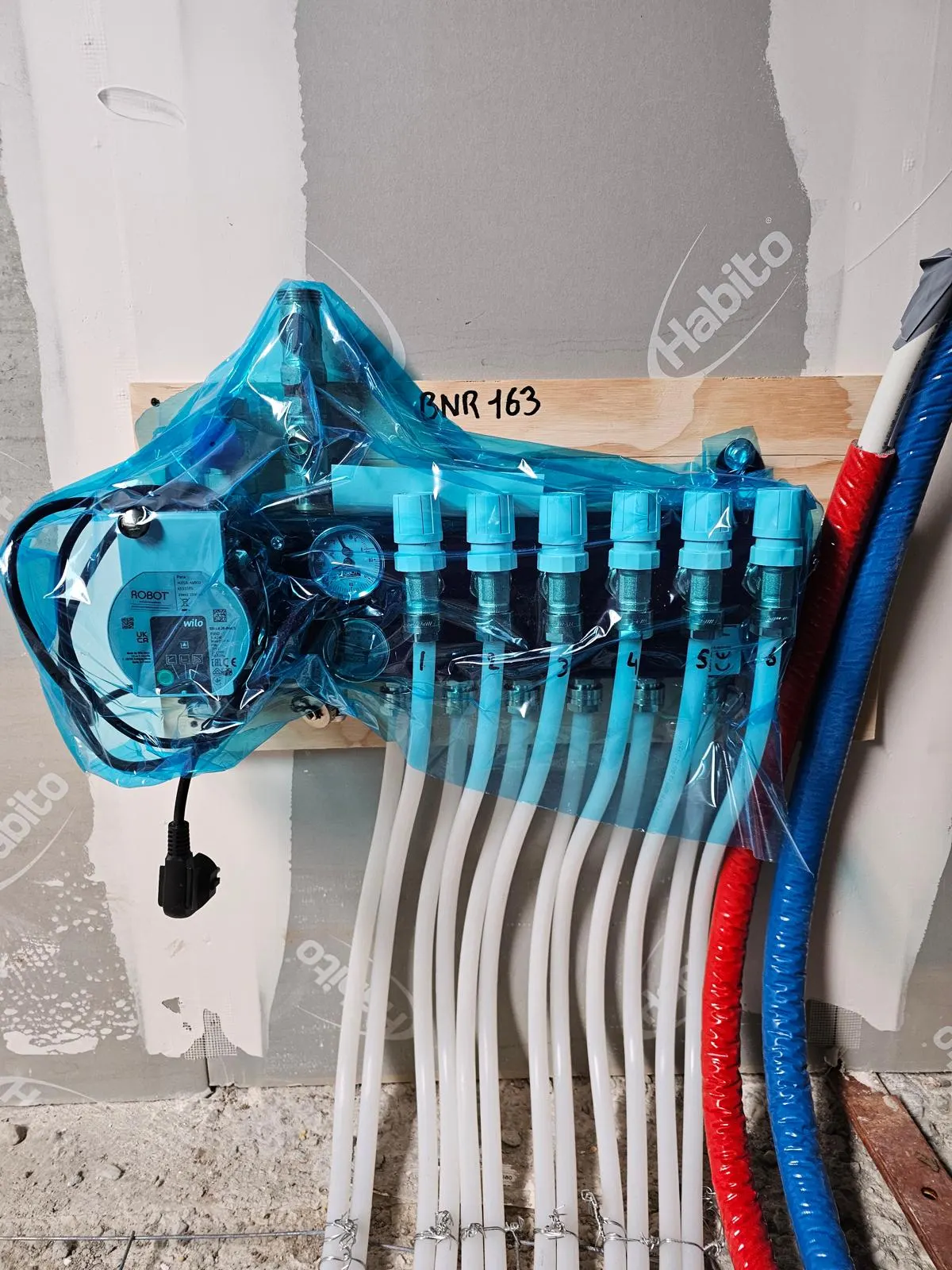 Een verdeler met zes groepen in blauwe beschermfolie op een houten montageplaat tegen een gipswand, met de pomp links en de rode en blauwe geïsoleerde aanvoerleidingen rechts ernaast