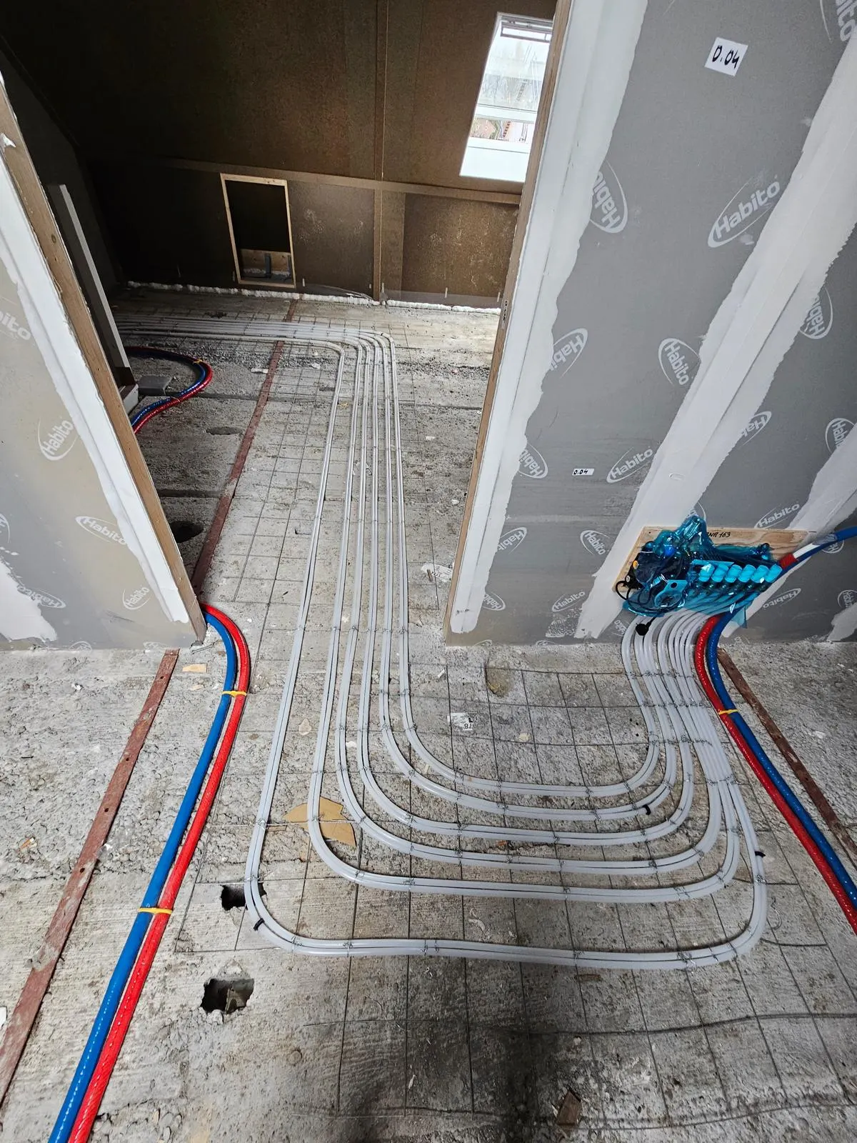 Bundels vloerverwarmingsbuis die door een deuropening van de ene ruimte naar de andere lopen over een wapeningsnet op een kale betonvloer, met rode en blauwe aanvoerleidingen langs de wand