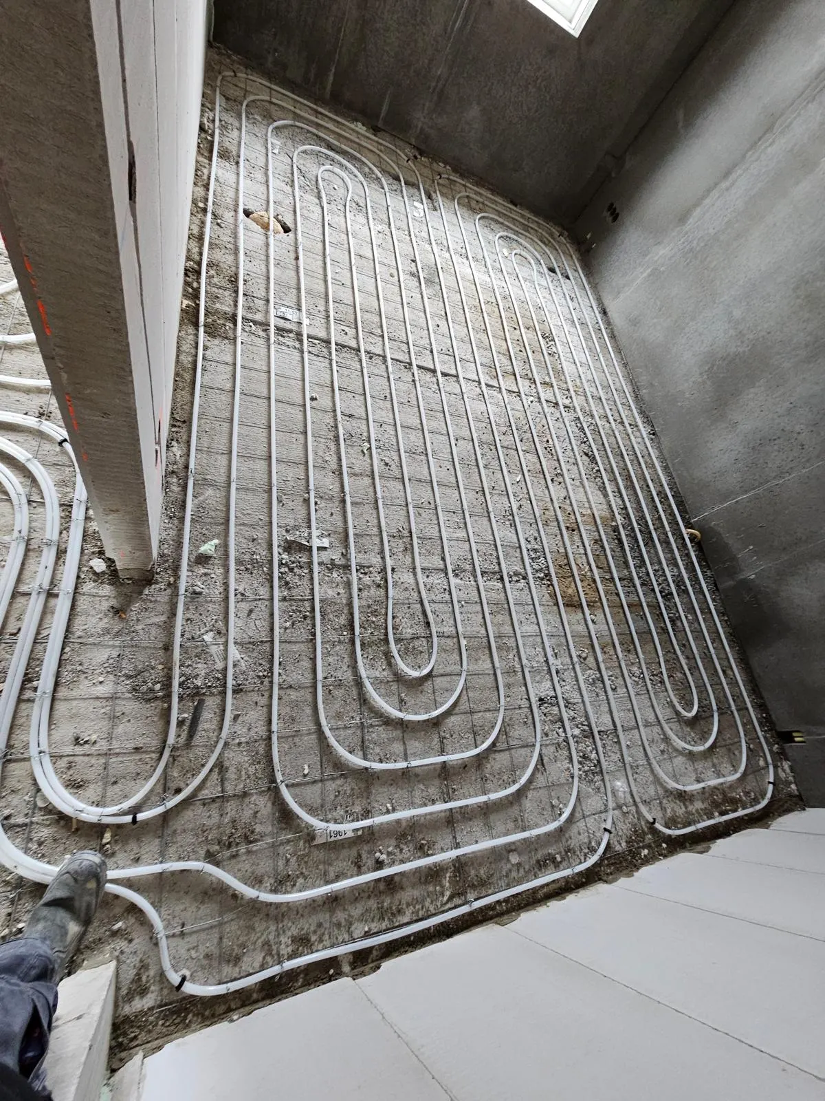 Een vlak vol vloerverwarmingsbuis op een wapeningsnet langs een betonnen wand, met de gestorte dekvloer rechts ernaast