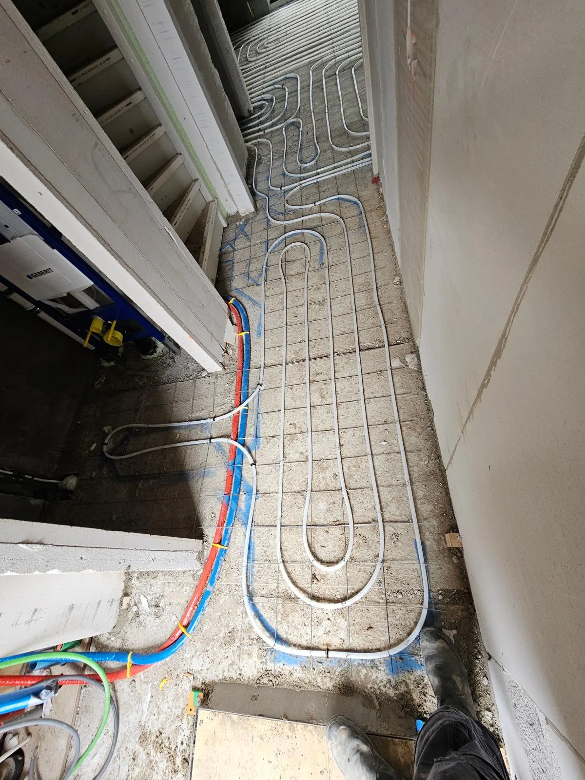 Vloerverwarmingsbuis in lange smalle lussen op een wapeningsnet in een kleine ruimte, met blauwe uitzetmarkeringen op de vloer en de aanvoerleidingen langs de wand