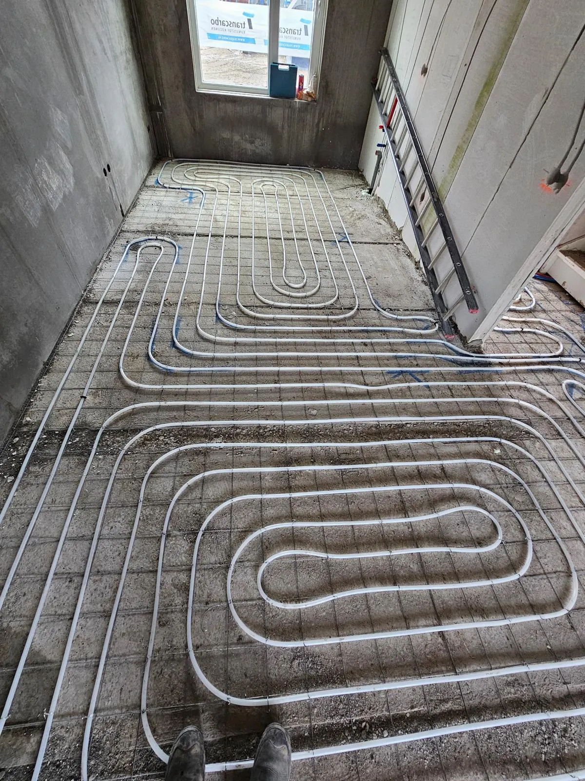 Een ruimte met betonwanden vol vloerverwarmingsbuis in brede lussen op een wapeningsnet, met een ladder tegen de wand en daglicht uit een raam op de achtergrond