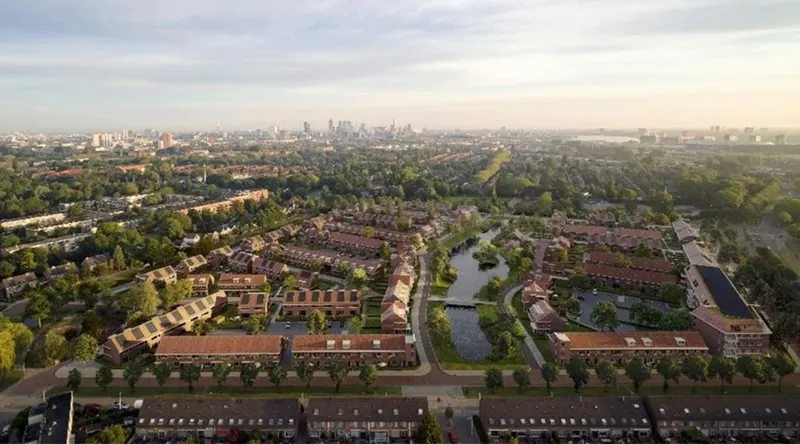 Een luchtopname van een nieuwbouwwijk met rijen grondgebonden woningen in rode baksteen met kappen en zonnepanelen, rond een gegraven watergang met een eilandje en jonge beplanting, met bomenrijen langs de randen en de skyline van Rotterdam op de achtergrond