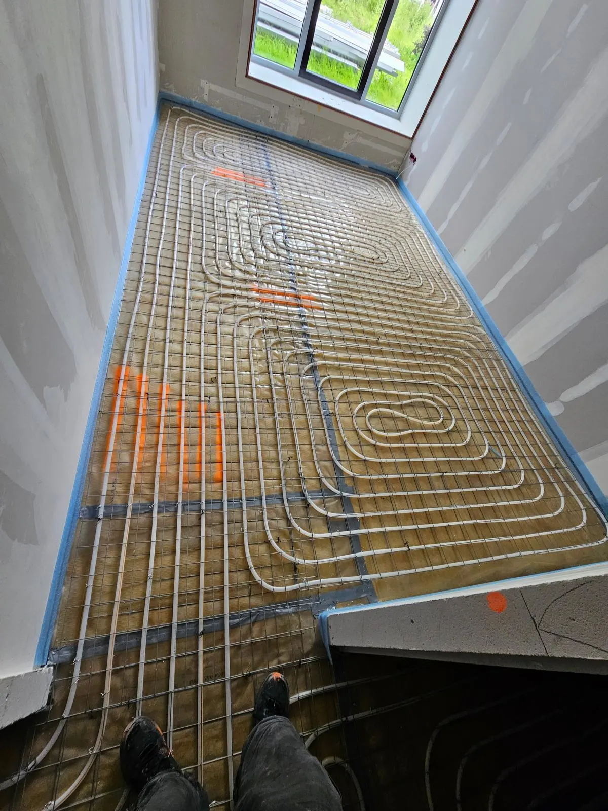 Een smalle kamer met een raam aan de kopse kant, met vloerverwarmingsbuis in lange lussen en een klein slakkenhuis in het midden, op een wapeningsnet over lichtbruine plaatisolatie met blauwe randisolatie langs de wanden