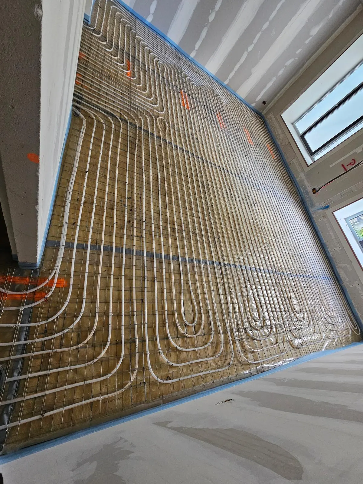 Vloerverwarmingsbuis die in dichte banen over een kamervloer loopt en bij de wand in ruime bochten terugdraait, op een wapeningsnet over lichtbruine plaatisolatie, met gipsplaatwanden en twee ramen op de achtergrond