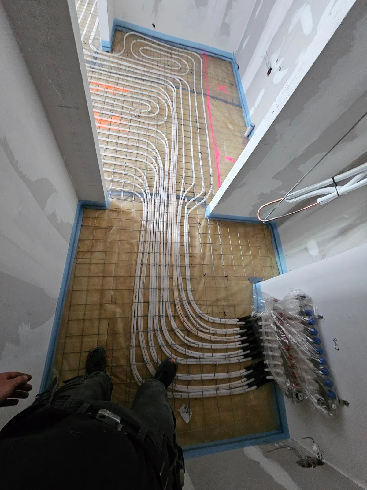 Een kamer met een verdeler in beschermfolie aan de wand rechts, waar de witte leidingen in zwarte bochtsteunen naar beneden komen en over het wapeningsnet de ruimte in lopen