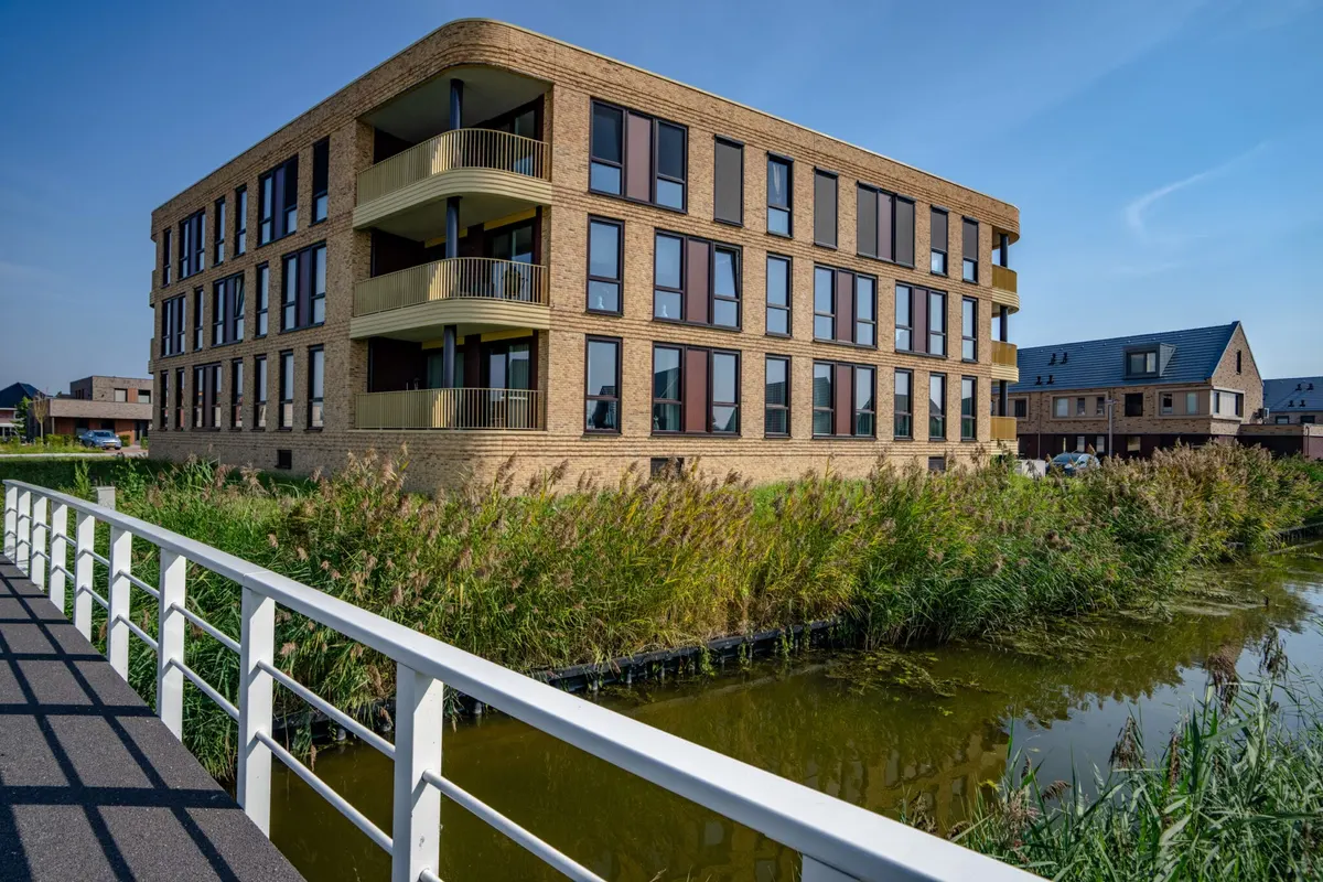 Een opgeleverd appartementengebouw van drie lagen in geelbruine baksteen met een afgeronde hoek, met drie balkons boven elkaar met goudkleurige spijlen, aan een sloot met riet langs de oever en een witte brugleuning op de voorgrond