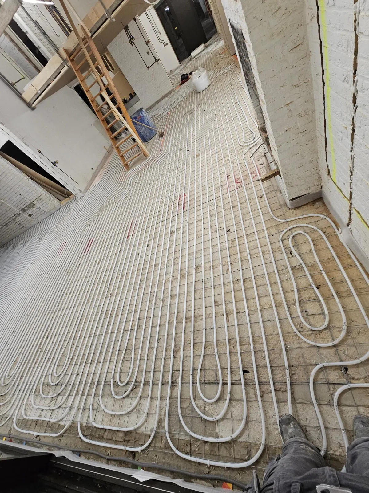 Lange banen vloerverwarmingsbuis op een wapeningsnet over de volle diepte van een villa in verbouwing, met witgeschilderd metselwerk langs de wanden