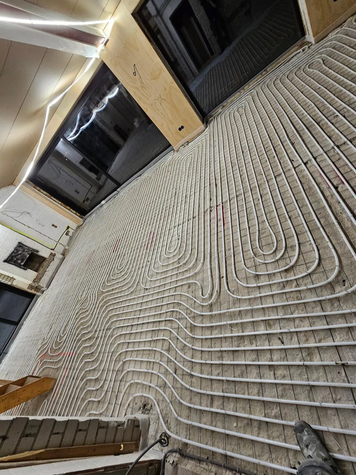 Vloerverwarmingsbuis in dichte lussen op een wapeningsnet onder een verlaagd plafond met een houten balk, met daglicht uit twee hoge ramen