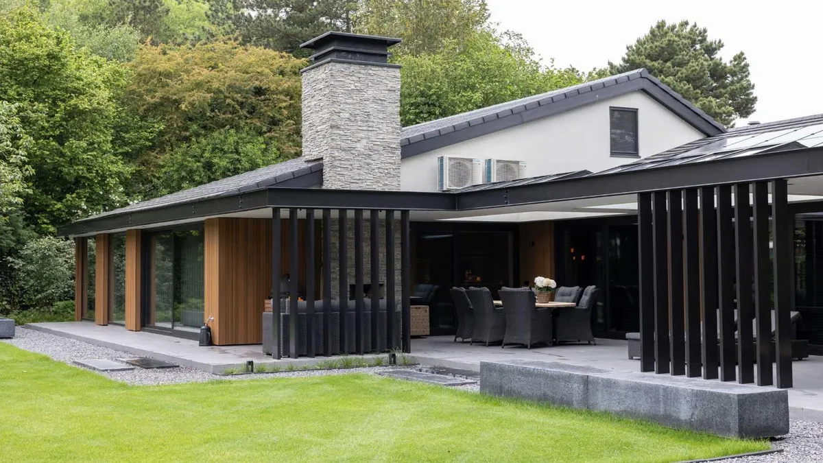Een opgeleverde vrijstaande villa gezien vanaf het gazon, met lage ver overstekende daken met zwarte randen, een schoorsteen van gestapelde natuursteen, gevelbekleding in verticaal houten latwerk naast schuifpuien over de volle hoogte, en rechts een terrasoverkapping op zwarte staanders met een eettafel eronder